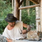 Ein Mann mit Hut und weißem Hemd bearbeitet einen Stein mit Hammer und Meißel in der Nähe einer Holzkonstruktion.