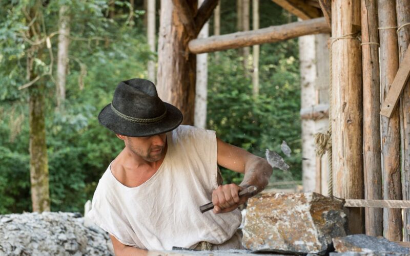 Ein Mann mit Hut und weißem Hemd bearbeitet einen Stein mit Hammer und Meißel in der Nähe einer Holzkonstruktion.
