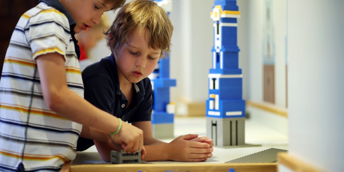 Zwei Jungen bauen mit blauen und weißen Lego-Steinen an einem Tisch, im Hintergrund sind hohe Lego-Strukturen zu sehen.