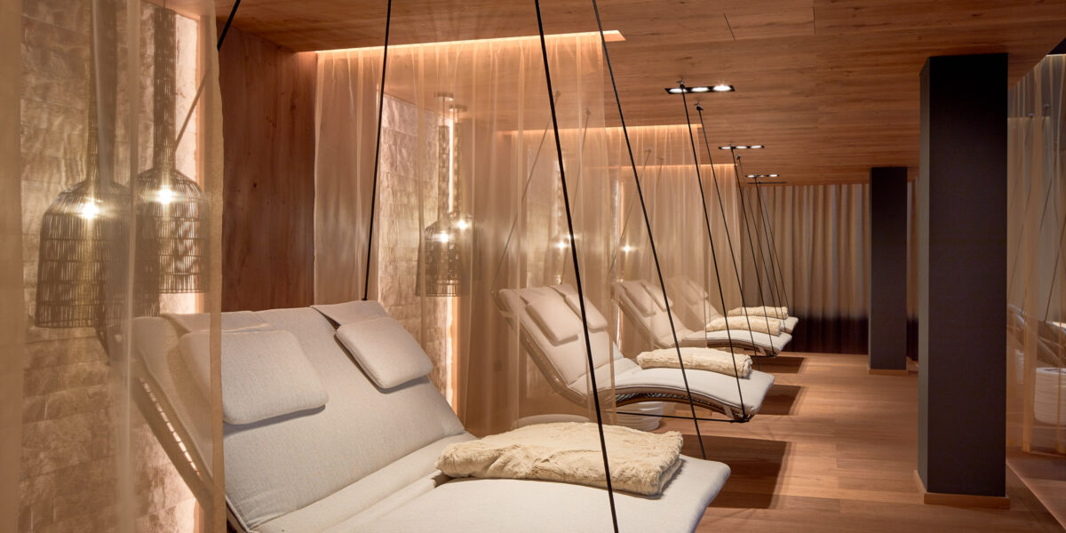 Moderne Entspannungslounge mit gepolsterten Schaukelstühlen, gedämpfter Beleuchtung, Holzböden und -decken sowie halbtransparenten Vorhängen, die jeden Sitzbereich abtrennen.