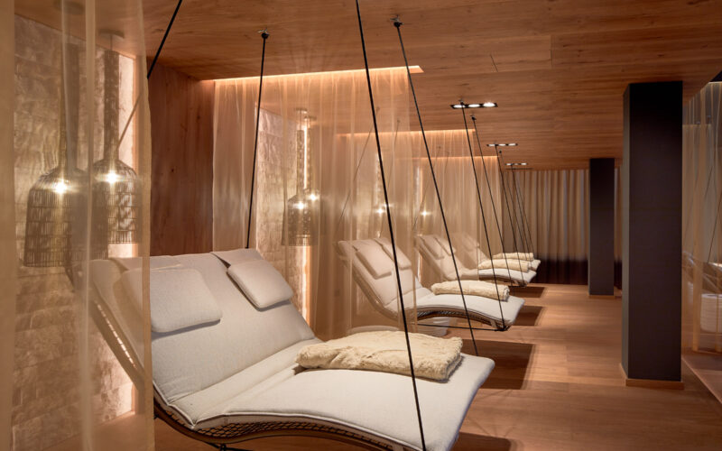 Moderne Entspannungslounge mit gepolsterten Schaukelstühlen, gedämpfter Beleuchtung, Holzböden und -decken sowie halbtransparenten Vorhängen, die jeden Sitzbereich abtrennen.