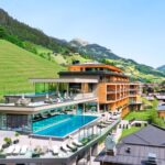 Modernes, mehrstöckiges Resort mit Außenpools und Lounge-Bereichen, das auf einem Hügel mit grünen Hängen und Bergen im Hintergrund unter einem klaren blauen Himmel liegt.
