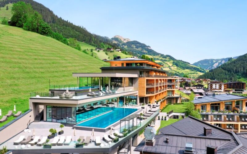 Modernes, mehrstöckiges Resort mit Außenpools und Lounge-Bereichen, das auf einem Hügel mit grünen Hängen und Bergen im Hintergrund unter einem klaren blauen Himmel liegt.