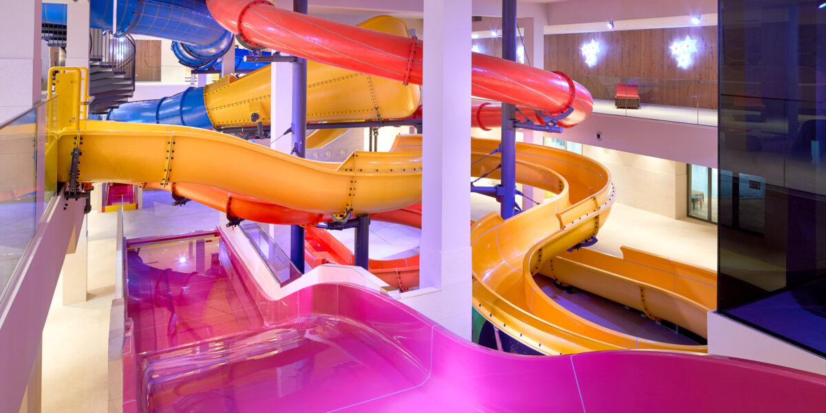 Farbenfrohe Indoor-Wasserrutschen mit gelben, rosa, roten und blauen Röhren, die sich um weiße Säulen in einer gut beleuchteten Freizeitanlage winden.