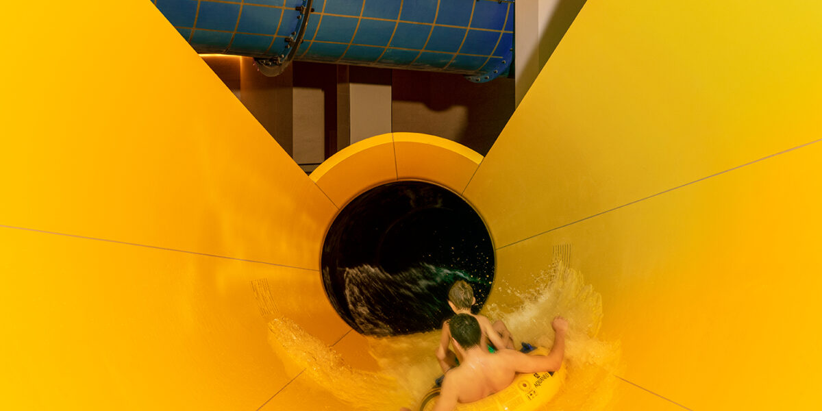 Zwei Kinder fahren auf einem gelben Schlauchboot eine bunte Wasserrutsche hinunter und nähern sich einem dunklen Tunnelabschnitt.