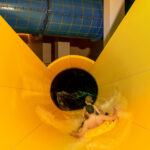 Zwei Kinder fahren auf einem gelben Schlauchboot eine bunte Wasserrutsche hinunter und nähern sich einem dunklen Tunnelabschnitt.