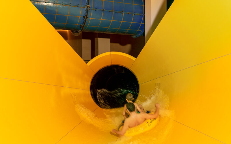 Zwei Kinder fahren auf einem gelben Schlauchboot eine bunte Wasserrutsche hinunter und nähern sich einem dunklen Tunnelabschnitt.