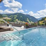Außenschwimmbad auf einer Terrasse mit Liegestühlen und Blick auf die Berge unter einem teilweise bewölkten Himmel.