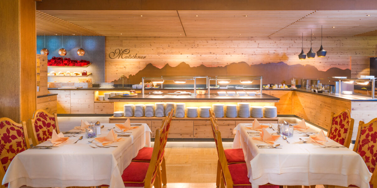 Ein Restaurant mit gedeckten Tischen, rot-goldenen Stühlen und einer Buffet-Theke mit Tellern und Speisenauslagen im Hintergrund.