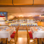 Ein Restaurant mit gedeckten Tischen, rot-goldenen Stühlen und einer Buffet-Theke mit Tellern und Speisenauslagen im Hintergrund.