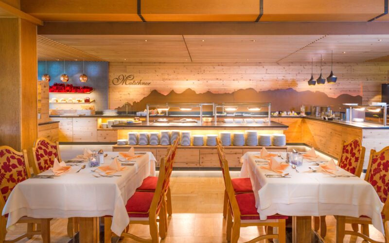 Ein Restaurant mit gedeckten Tischen, rot-goldenen Stühlen und einer Buffet-Theke mit Tellern und Speisenauslagen im Hintergrund.