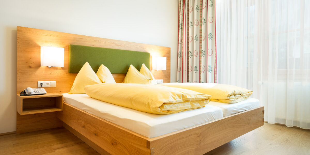Ein modernes Hotelzimmer mit einem hölzernen Doppelbett, einem grünen Kopfteil, gelber Bettwäsche, zwei Wandlampen, einem Beistelltisch mit einem Telefon und einem Fenster mit durchsichtigen und geblümten Vorhängen.