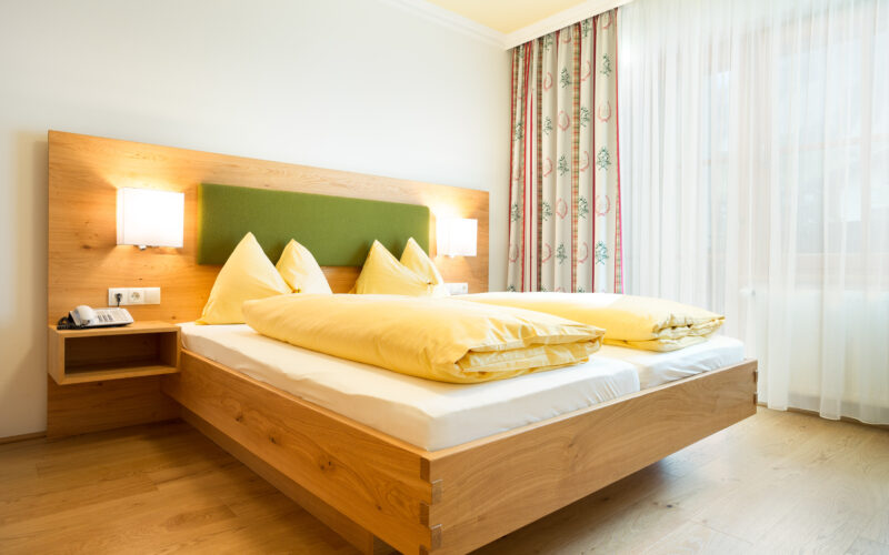 Ein modernes Hotelzimmer mit einem hölzernen Doppelbett, einem grünen Kopfteil, gelber Bettwäsche, zwei Wandlampen, einem Beistelltisch mit einem Telefon und einem Fenster mit durchsichtigen und geblümten Vorhängen.