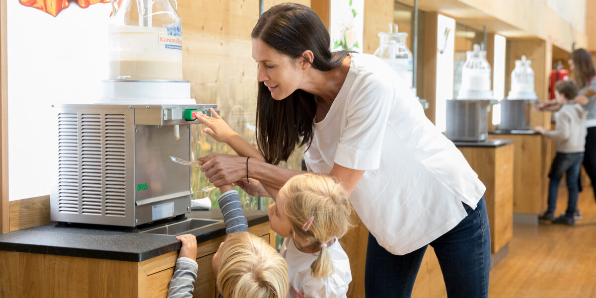 Eine Frau hilft zwei kleinen Kindern, einen Milchspender in einem hellen Innenraum mit Holzböden und Displays zu bedienen.