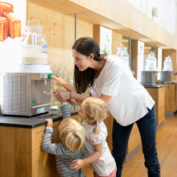 Eine Frau hilft zwei kleinen Kindern, einen Milchspender in einem hellen Innenraum mit Holzböden und Displays zu bedienen.