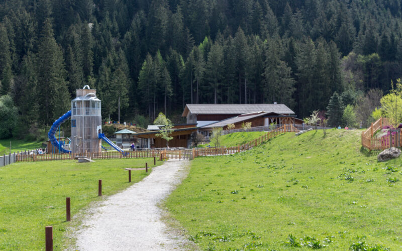 Ein Schotterweg führt zu einem Spielplatz mit einer blauen Rutsche und einem Kletterturm, der auf einer Rasenfläche in der Nähe eines bewaldeten Hügels liegt.