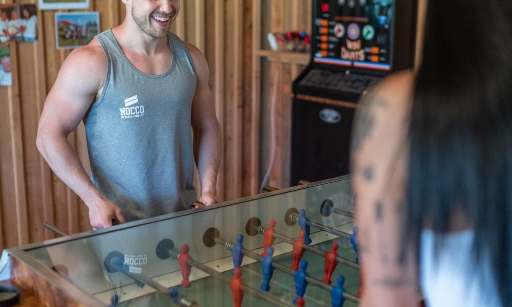 Zwei Personen spielen in einem Holzzimmer Tischfußball. Ein Mann in einem Tank-Top lächelt, während eine Frau mit langen schwarzen Haaren ihm gegenüber steht. Im Hintergrund steht ein Dartboard-Automat.