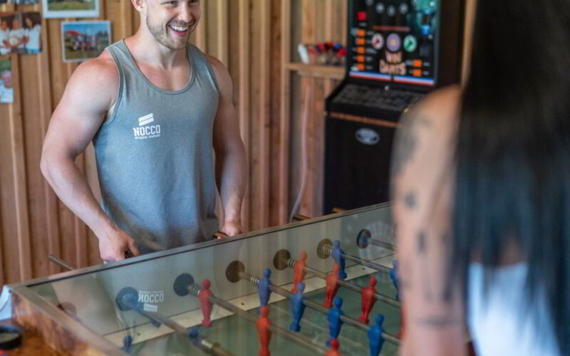 Zwei Personen spielen in einem Holzzimmer Tischfußball. Ein Mann in einem Tank-Top lächelt, während eine Frau mit langen schwarzen Haaren ihm gegenüber steht. Im Hintergrund steht ein Dartboard-Automat.