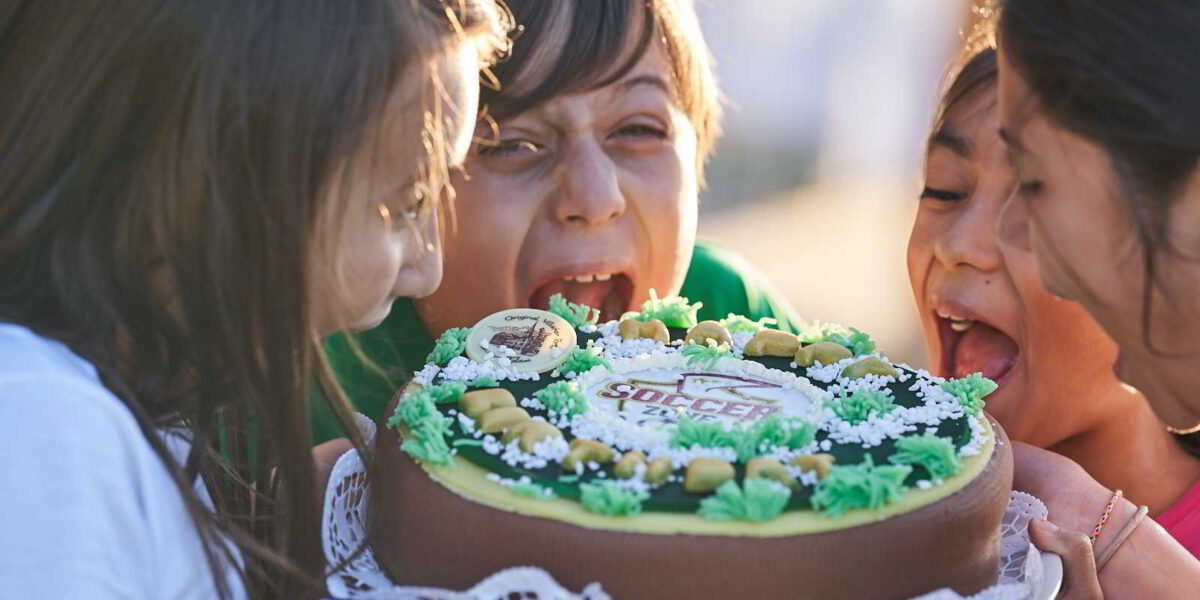 Vier Kinder essen eifrig einen dekorierten Kuchen und lecken ihn gemeinsam ab. Sie wirken fröhlich und verspielt, während sie um das Dessert versammelt sind.