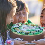 Vier Kinder essen eifrig einen dekorierten Kuchen und lecken ihn gemeinsam ab. Sie wirken fröhlich und verspielt, während sie um das Dessert versammelt sind.