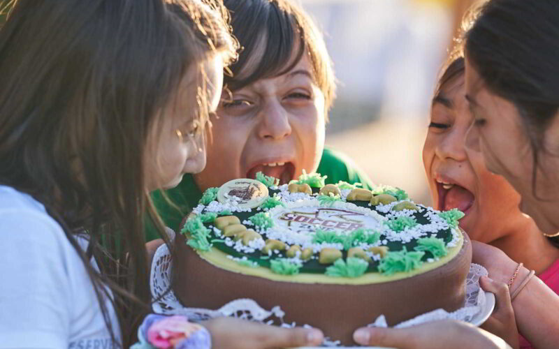 Vier Kinder essen eifrig einen dekorierten Kuchen und lecken ihn gemeinsam ab. Sie wirken fröhlich und verspielt, während sie um das Dessert versammelt sind.
