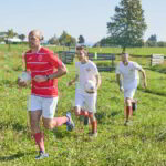 Drei Männer, einer in einem roten Sportdress und zwei in Weiß, laufen an einem sonnigen Tag mit Fußbällen in der Hand über eine Wiese.