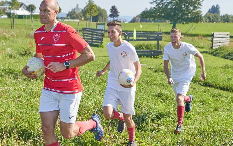 Drei Männer, einer in einem roten Sportdress und zwei in Weiß, laufen an einem sonnigen Tag mit Fußbällen in der Hand über eine Wiese.