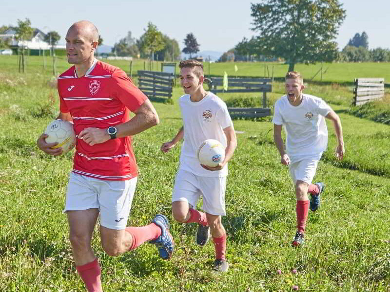 Drei Männer, einer in einem roten Sportdress und zwei in Weiß, laufen an einem sonnigen Tag mit Fußbällen in der Hand über eine Wiese.