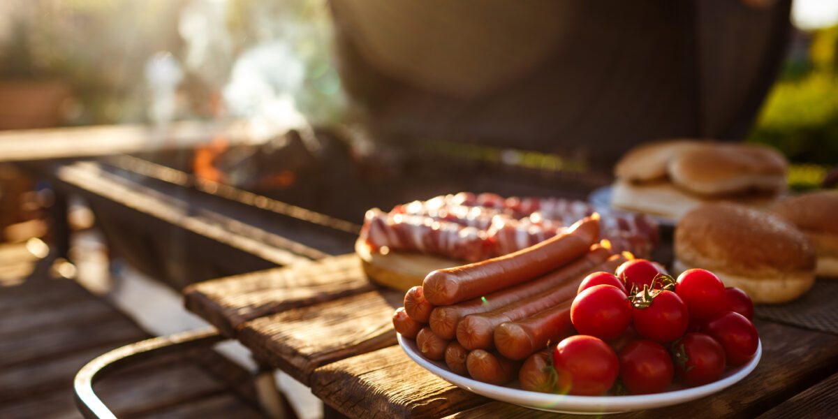 Ein Teller mit rohen Hot Dogs und Kirschtomaten auf einem Holztisch neben einem Grill mit Würstchen, Brötchen und Rauch im Hintergrund.