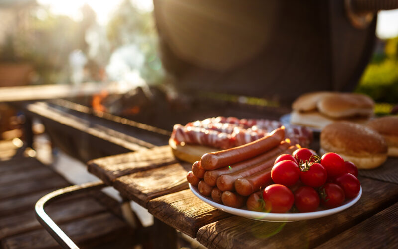 Ein Teller mit rohen Hot Dogs und Kirschtomaten auf einem Holztisch neben einem Grill mit Würstchen, Brötchen und Rauch im Hintergrund.