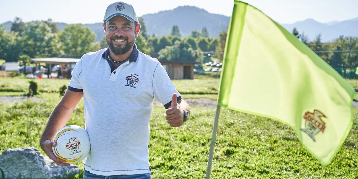 Ein Mann in weißem Polohemd und Mütze steht im Freien, hält einen Fußball in der Hand und zeigt den Daumen nach oben neben einer leuchtend grünen Fahne auf einer Wiese.