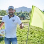 Ein Mann in weißem Polohemd und Mütze steht im Freien, hält einen Fußball in der Hand und zeigt den Daumen nach oben neben einer leuchtend grünen Fahne auf einer Wiese.