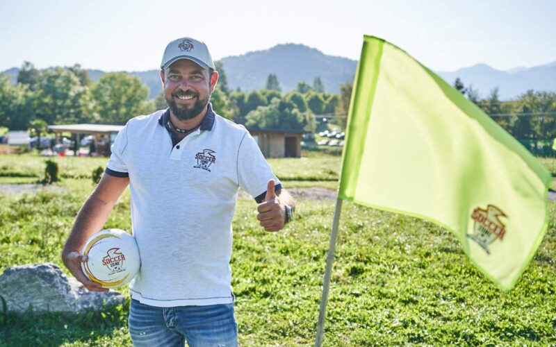 Ein Mann in weißem Polohemd und Mütze steht im Freien, hält einen Fußball in der Hand und zeigt den Daumen nach oben neben einer leuchtend grünen Fahne auf einer Wiese.