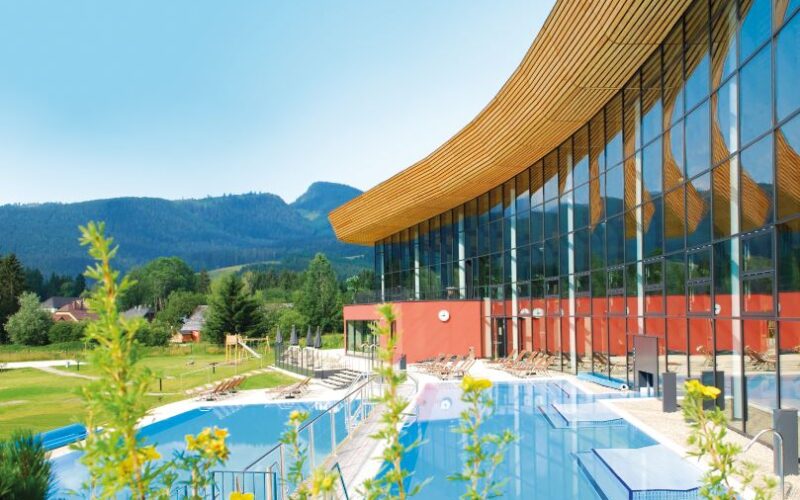 Modernes Gebäude mit großen Glasfenstern neben Außenpools, umgeben von grünen Rasenflächen und fernen Bergen unter einem klaren blauen Himmel.