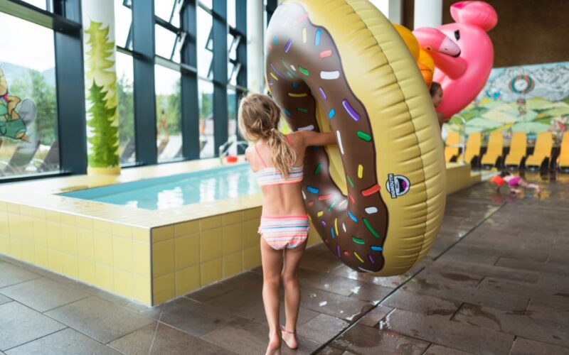Ein Kind im Badeanzug steht drinnen an einem kleinen Pool und hält einen großen aufblasbaren Donut-Schwimmer mit Streuseln und einen rosa Flamingo-Schwimmer dahinter.