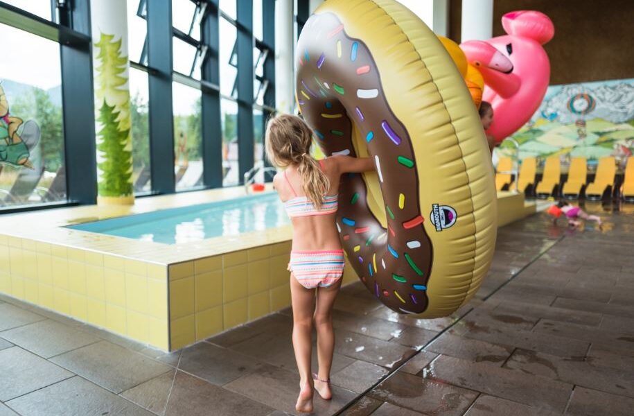 Ein Kind im Badeanzug steht drinnen an einem kleinen Pool und hält einen großen aufblasbaren Donut-Schwimmer mit Streuseln und einen rosa Flamingo-Schwimmer dahinter.