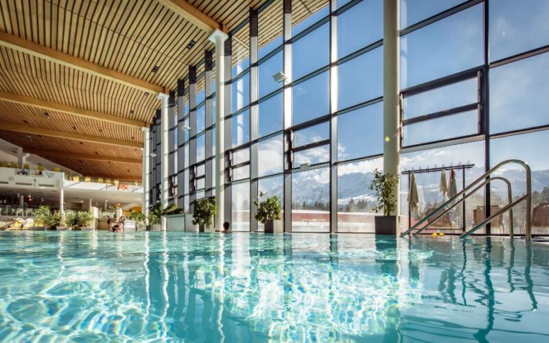 Hallenbad mit klarem Wasser, große, raumhohe Fenster mit Blick auf die schneebedeckten Berge und Holzdeckenpaneele. Topfpflanzen säumen die Fenster.