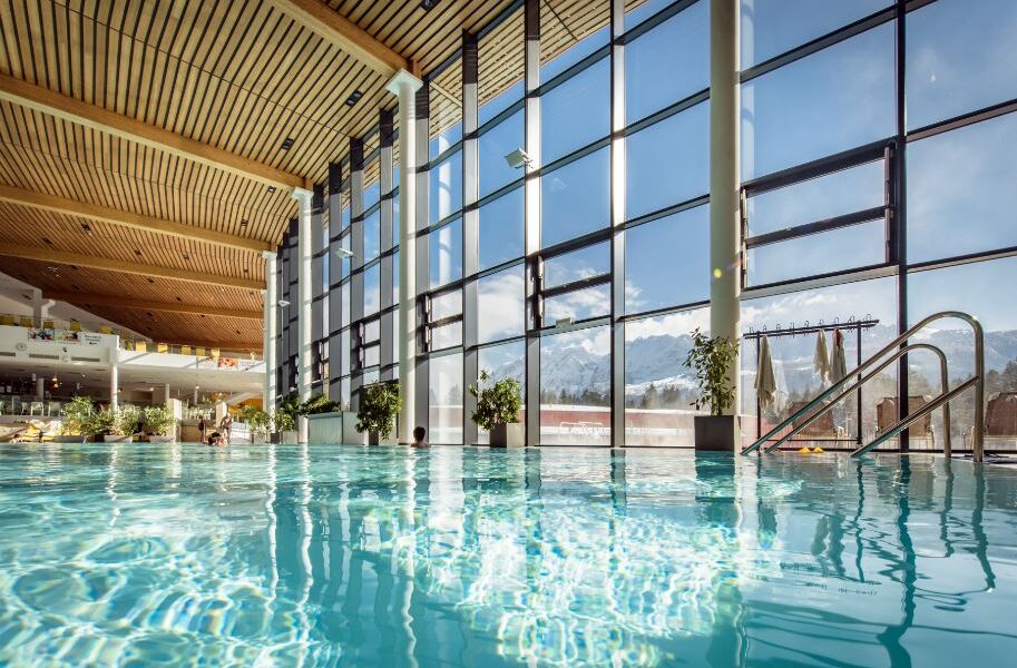 Hallenbad mit klarem Wasser, große, raumhohe Fenster mit Blick auf die schneebedeckten Berge und Holzdeckenpaneele. Topfpflanzen säumen die Fenster.