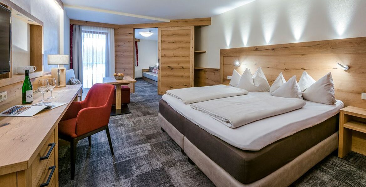 Modernes Hotelzimmer mit einem großen Doppelbett, Holzmöbeln, einem Schreibtisch mit rotem Stuhl, einer Sitzecke und einem Fenster mit durchsichtigen Vorhängen, durch das Tageslicht fällt.