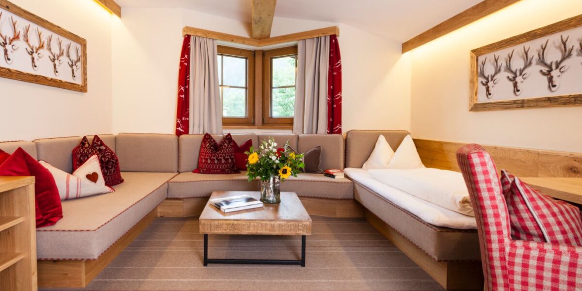Ein gemütliches Wohnzimmer mit einem Ecksofa, einem hölzernen Couchtisch mit einer Blumenvase, einem Schlafsofa und einer Einrichtung mit roten Akzenten und einem Hirschkopf-Kunstwerk.
