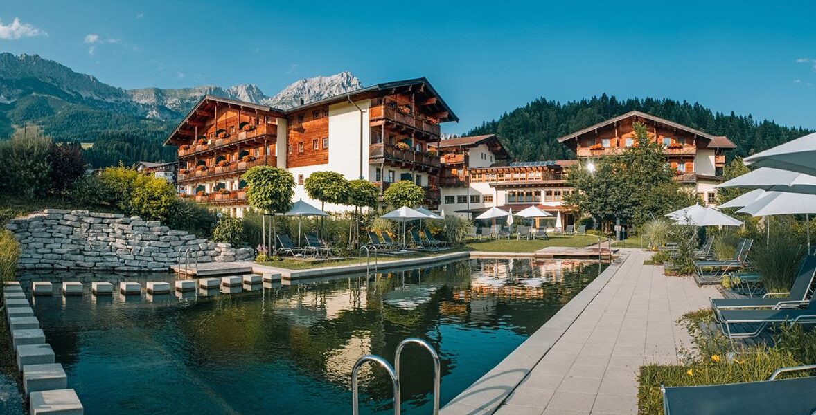 Ein Hotel mit Holzbalkonen liegt an einem angelegten Naturschwimmteich, umgeben von Liegestühlen, Sonnenschirmen und den Bergen im Hintergrund.