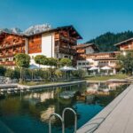 Ein Hotel mit Holzbalkonen liegt an einem angelegten Naturschwimmteich, umgeben von Liegestühlen, Sonnenschirmen und den Bergen im Hintergrund.