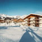 Die schneebedeckten Berghütten mit Holzbalkonen stehen vor der Kulisse der Berge unter einem strahlend blauen Himmel.