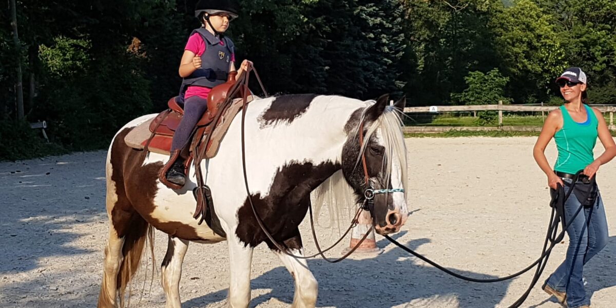 Ein junges Mädchen mit Helm reitet auf einem schwarz-weißen Pferd, während eine erwachsene Frau in einem Tank-Top das Pferd an einem sonnigen Tag führt.