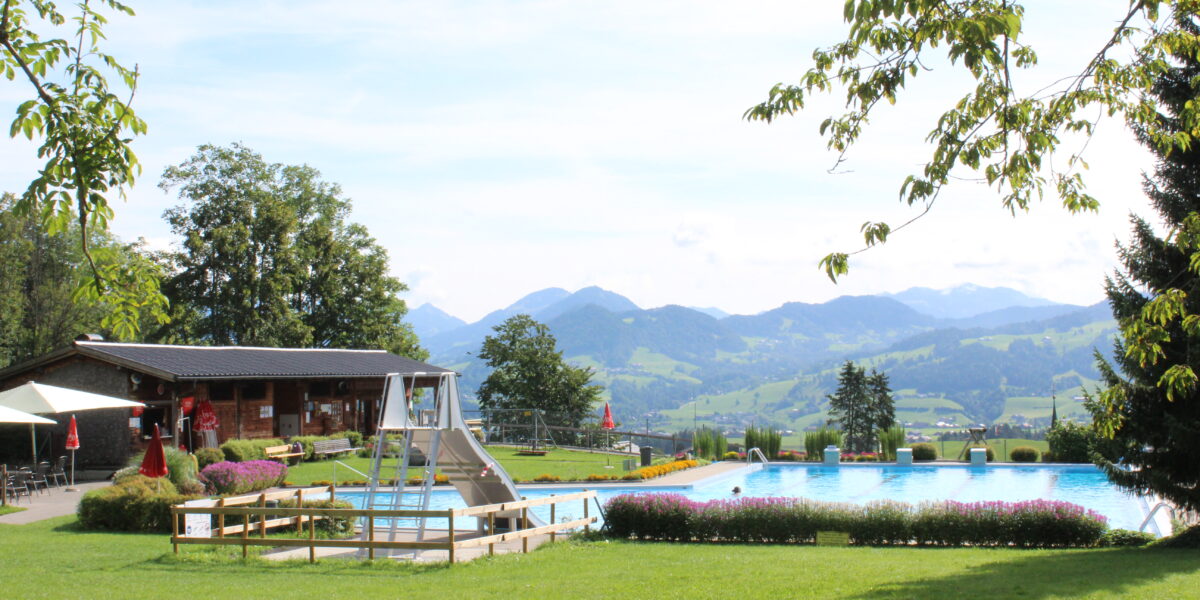 Außenschwimmbad mit einem Holzgebäude in der Nähe, umgeben von grünem Gras und Bäumen, mit Bergen im Hintergrund unter einem teilweise bewölkten Himmel.