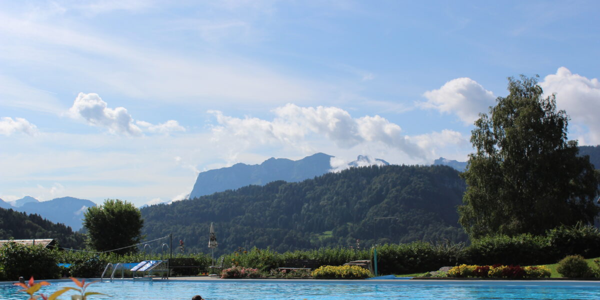 Außenschwimmbad mit einer Person im Wasser, umgeben von Grün und Bergen unter einem blauen Himmel mit vereinzelten Wolken.