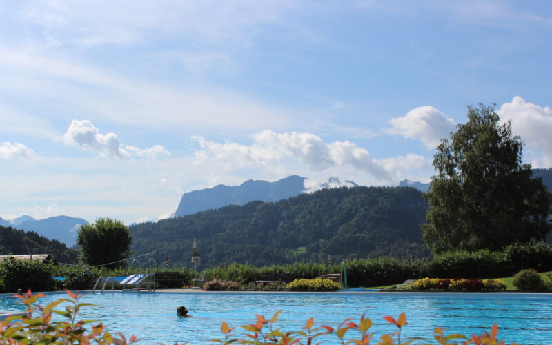 Außenschwimmbad mit einer Person im Wasser, umgeben von Grün und Bergen unter einem blauen Himmel mit vereinzelten Wolken.