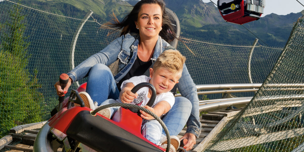 Eine Frau und ein kleiner Junge fahren mit einer roten Alpenachterbahn eine Strecke hinunter, auf der Seilbahnen und Berge im Hintergrund zu sehen sind.