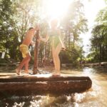 Zwei Kinder in Badeanzügen stehen auf einem Holzfloß in einem sonnenbeschienenen, flachen, von Bäumen umgebenen Teich.