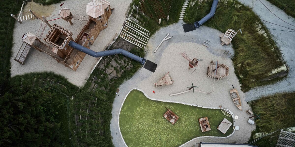 Luftaufnahme eines Spielplatzes mit Holzkonstruktionen, Rutschen, Klettergerüsten, Kieswegen und einem kleinen grünen Teich, der von Gras und Vegetation umgeben ist.